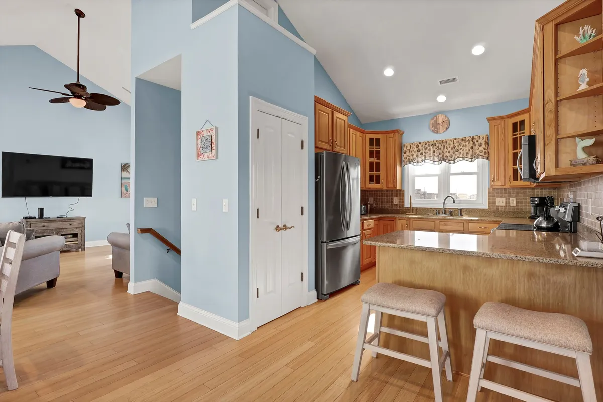 A spacious, open-plan living area featuring a kitchen island with seating, modern kitchen appliances, a cozy living room area with plush seating, and a TV mounted on the wall. Natural light fills the room, creating a bright, inviting atmosphere ideal for gatherings.