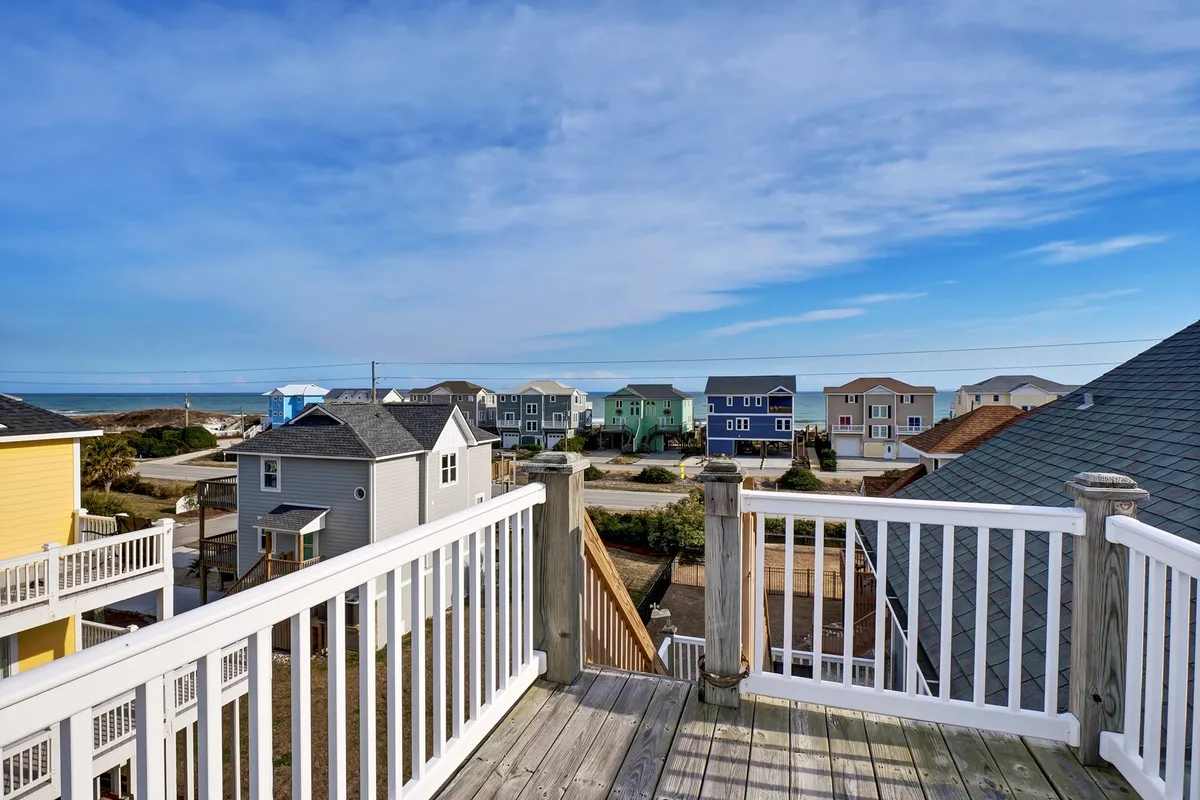 A cheerful balcony scene showing a railing overlooking the ocean and colorful neighboring houses. The balcony is positioned for relaxation, socializing, and enjoying expansive sunset views.