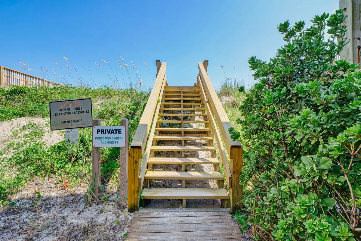 A wooden staircase flanked by lush greenery leads straight to a sandy beach. Signs mark the path, emphasizing private access for residents and guests. The morning light creates a serene, inviting mood for a day on the shore.