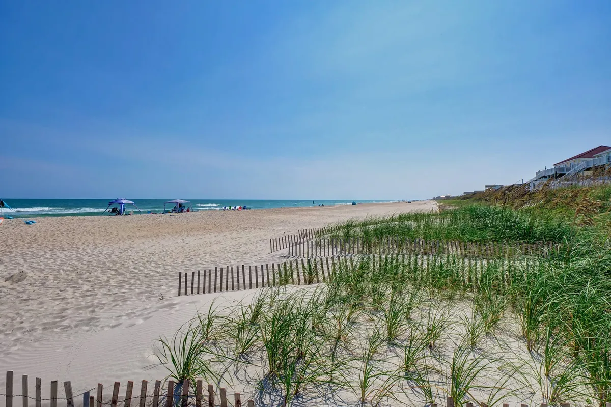 A sun-filled stretch of North Topsail Beach with soft sand, gentle ocean waves, and colorful beach umbrellas dotting the shoreline. Children and families are seen building sandcastles and playing beside the water, while the dunes provide a natural, scenic backdrop.