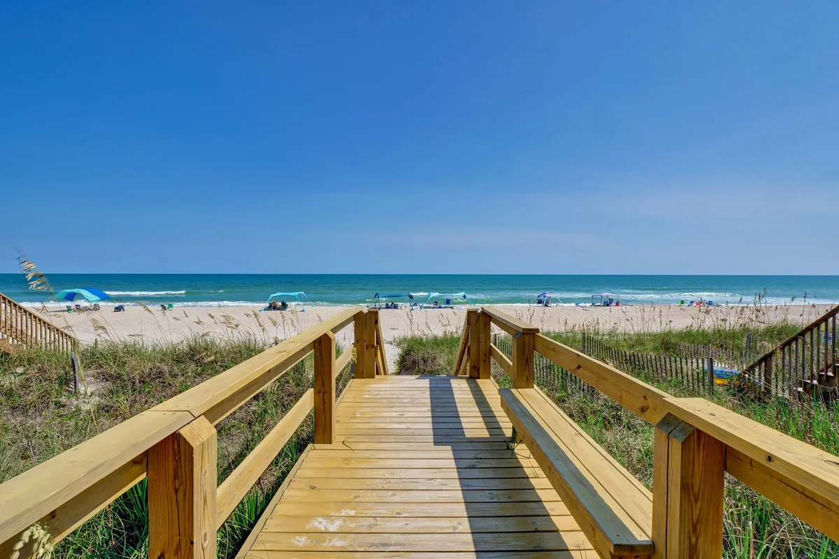A wide wooden boardwalk leads directly to the beach, where distant umbrellas and blue ocean water set the scene. The perspective evokes easy access from home to shore, inviting families to start their days by the water.