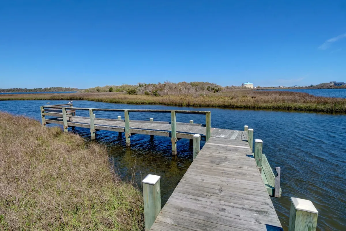 A tranquil scene featuring a community dock extending over calm water and surrounded by marshland. The wooden dock offers space for fishing, kayaking, or simply enjoying peaceful moments by the water. The setting is serene, inviting relaxation and connection with nature.