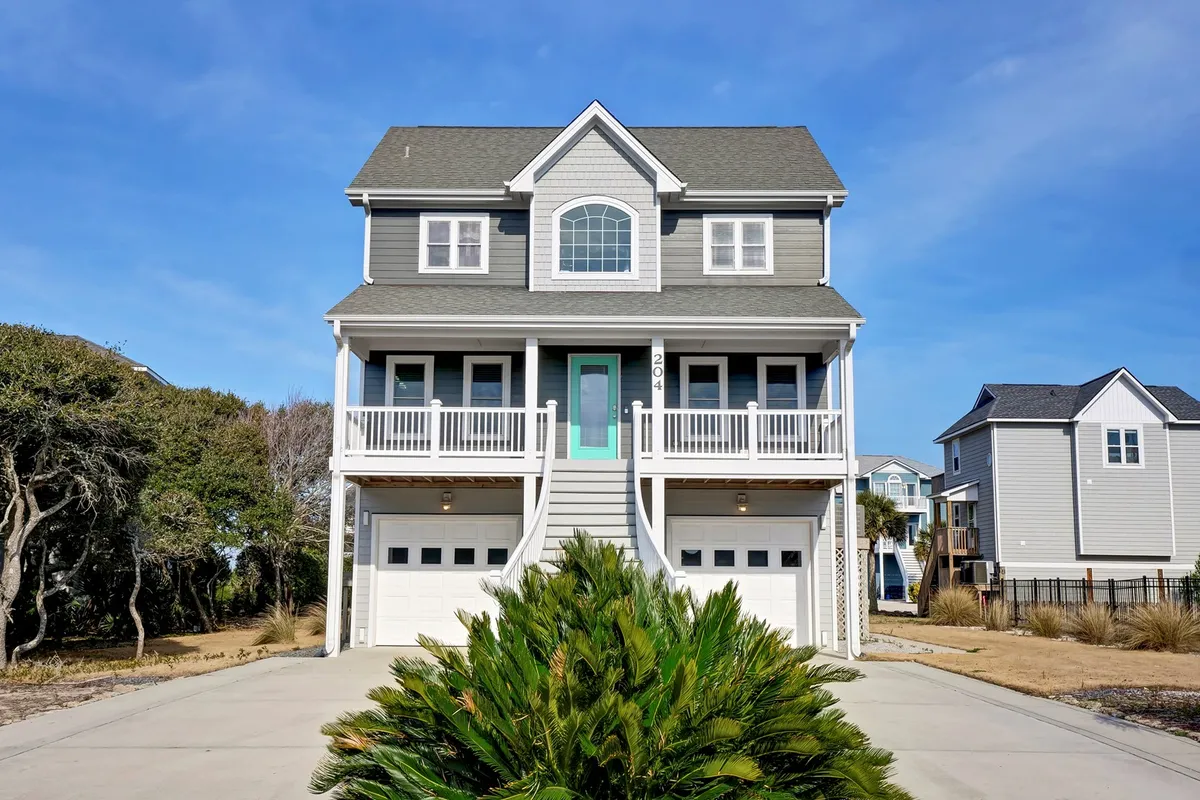The image showcases the inviting exterior of a single-family beach house, with a spacious front porch perfect for relaxing, a cheerful teal front door that adds a pop of color, and two garage doors hinting at plenty of room for guests. The atmosphere is bright and welcoming, with sunlight streaming onto the porch, evoking the promise of sunrise views, outdoor gatherings, and laid-back coastal living.