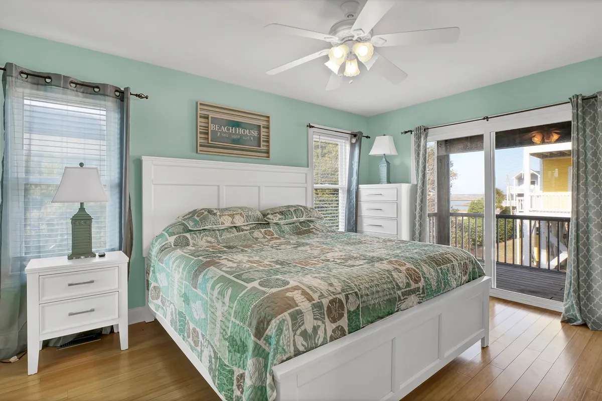 This interior image features a serene bedroom with a large bed, soft lamps, and a nightstand. A window allows sunlight to pour in, subtly hinting at an ocean view and the presence of a balcony just beyond. The overall mood is calm and restful, perfect for relaxation.