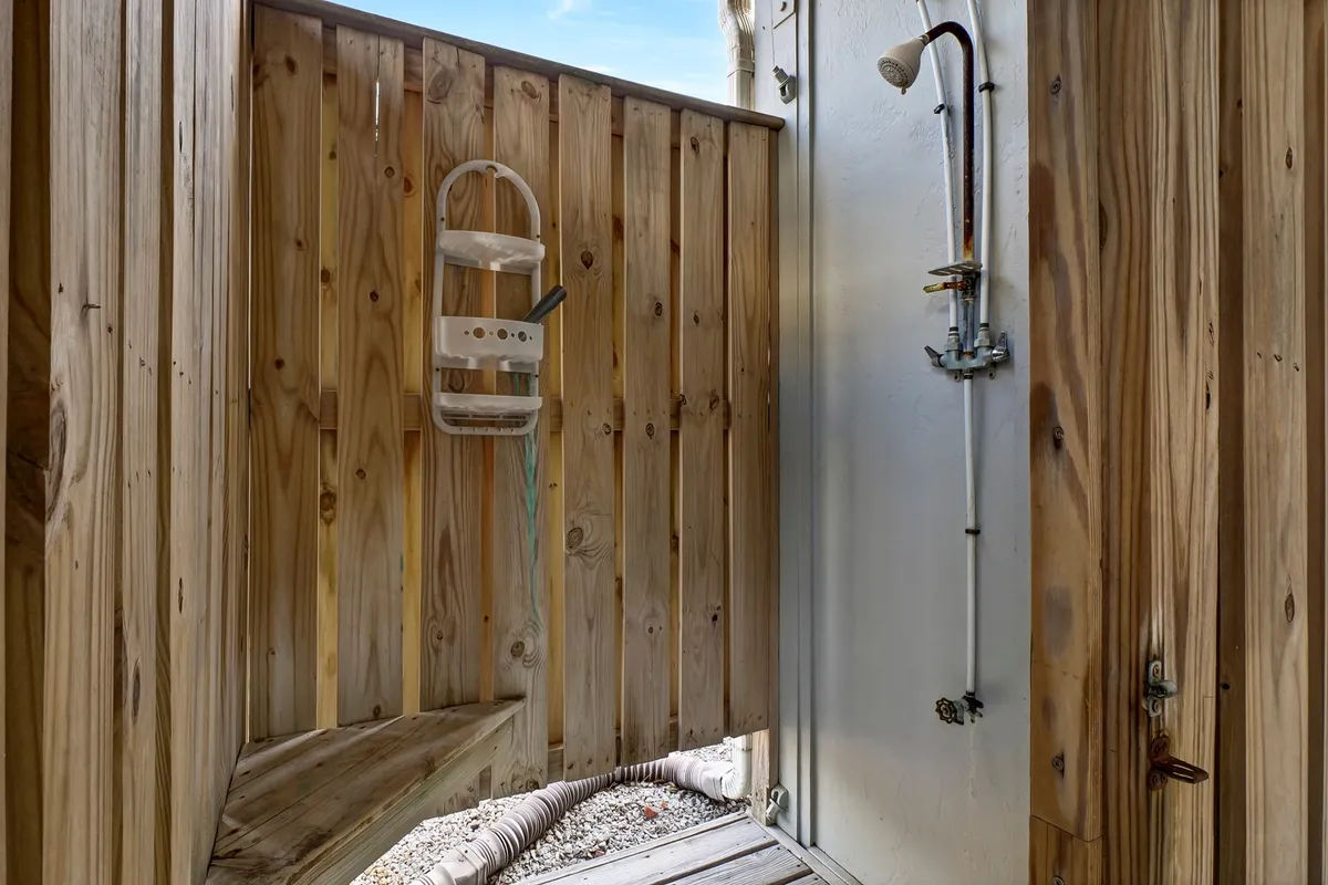 A charming exterior view of an outdoor shower with wooden walls, nestled in an open-air setting. Sunlight streams in, and the design highlights a practical amenity for washing off after beach adventures.